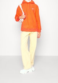 Felpa arancione con cappuccio e cordino, abbinata a pantaloni di velluto a coste leggeri giallo chiaro e scarpe platform bianche lucide.