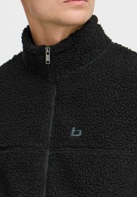 Chaqueta de forro polar negra con cuello alto, cierre frontal con cremallera y un logo sutil. La textura es suave y desaliñada con costuras visibles.