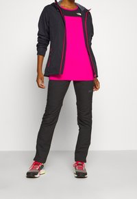 Schwarze leichte Jacke mit pinken Akzenten, kombiniert mit einem lebhaften pinken Hemd und schwarzen Hosen. Die Schuhe sind grau mit roten Schnürsenkeln und neonfarbenen Details.