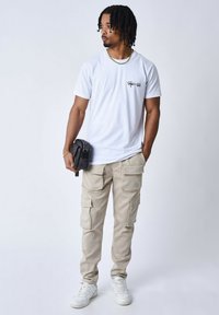 T-shirt bianca a girocollo con un piccolo logo nero, abbinata a pantaloni cargo beige chiaro con numerose tasche e sneakers bianche.