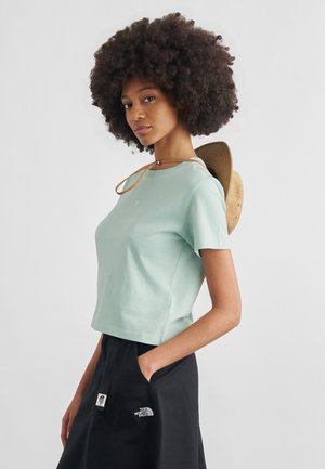 Žena s veľkými kučeravými vlasmi nosí svetlomodrý krátky rukáv, čiernu sukňu s logom The North Face a drží hnedý klobúk so širokou krempou za chrbtom.