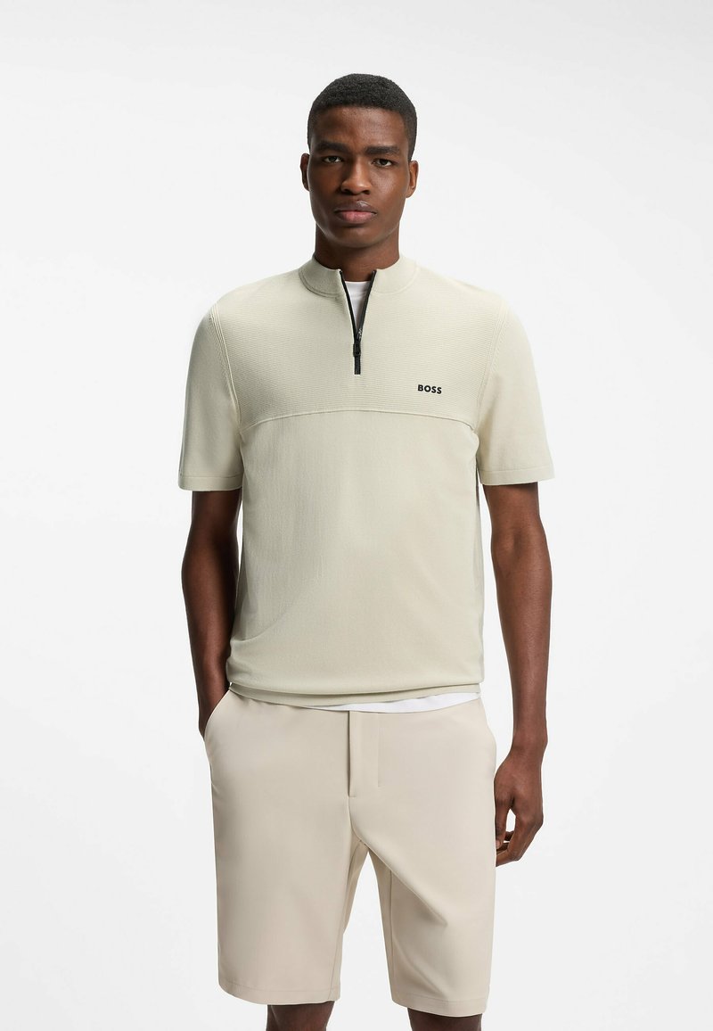 Homme portant une chemise zippée beige clair à manches courtes avec le logo "BOSS" et un short beige assorti, debout avec une main dans la poche.