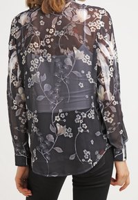 Transparente schwarze Bluse mit floralen Mustern in Weiß-, Creme- und sanften Farbtönen. Lange Ärmel, knöpfbare Manschetten und gerundeter Saum.