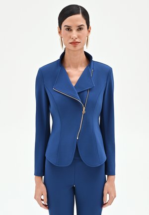 Femme aux cheveux noirs lisses portant une veste bleue cintrée avec une fermeture éclair dorée asymétrique et un pantalon bleu assorti sur un fond uni.