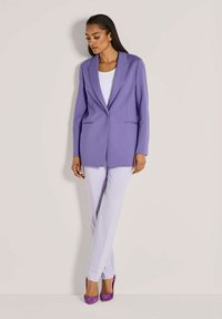 Vrouw die tegen een muur staat in een lila blazer, witte broek, witte top en paarse hoge hakken, naar beneden kijkend met handen ontspannen.