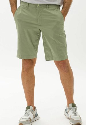 Mann trägt hellgrüne kniehohe Shorts, die Hände in den Taschen und weiße-graue Sneaker, steht vor einem weißen Hintergrund.