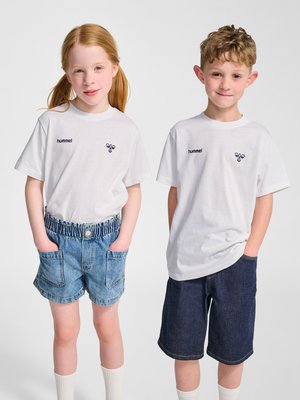 Deux enfants sont côte à côte, portant des t-shirts blancs hummel, des shorts en jean et des chaussettes blanches sur un fond uni.