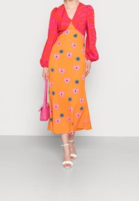 Longue robe orange avec des cœurs roses et des étoiles bleues, présentant un décolleté en V et des manches bouffantes, accompagnée de sandales blanches et d'un sac à main rose.