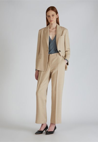 Beige op maat gemaakte pak met een blazer met één knoop en wijde broek, gecombineerd met een grijze V-hals top en zwarte puntige hakken.