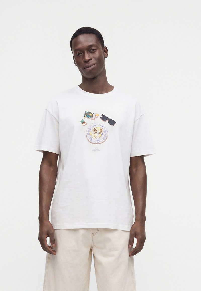 Άνδρας που φοράει λευκό t-shirt με εκτυπωμένα γυαλιά ηλίου, vintage τηλέφωνο, παγωτό και πιάτο με φαγητό, συνδυασμένα με ανοιχτό μπεζ παντελόνι.