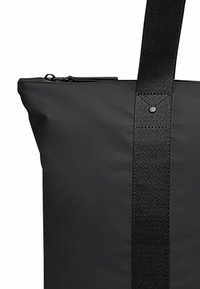 Svart toteveske med glatt ytterside, som har glidelås på toppen og bred svart stropp, preget av et rundt nagle og sydd detaljer.