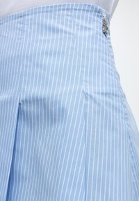 Φούστα ανοιχτού μπλε χρώματος με κάθετες λευκές ρίγες, πλισέ σχέδιο και ασημένιο κούμπωμα στο πλάι, φορεμένη πάνω από λευκό τοπ.