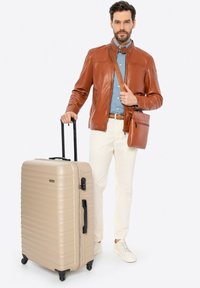 Valigia rigida beige, con texture a righe, zipper nera, ruote inline, tenuta da un uomo in giacca marrone e camicia blu.