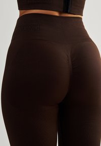 Hnědé legíny s vysokým pasem, ribbed texturou a bezšvovým designem. Mají logo na pase a hladký, přiléhavý siluetu.