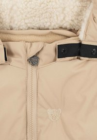 Chaqueta infantil color beige con un forro de felpa color crema. Cuenta con un tirador de cremallera en forma de oso y acentos en tiras negras.