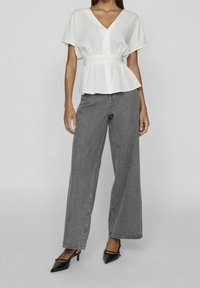Blusa bianca con peplum, maniche corte e scollo a V, abbinata a jeans grigi in denim larghi e scarpe col tacco nere in vernice.