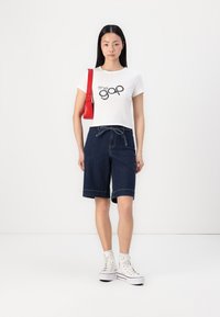 Biele tričko s logom "the gap", tmavomodré džínsy šortky a biele vysoké tenisky. Červená kabelka je prehodená cez rameno.