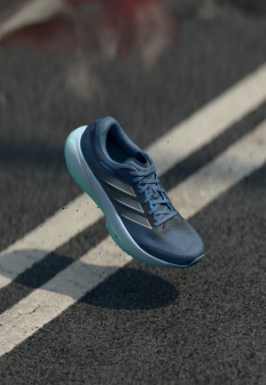 Niebieski but do biegania Adidas w powietrzu nad asfaltem z białymi oznaczeniami pasów, ukazujący podeszwę i boczne paski.