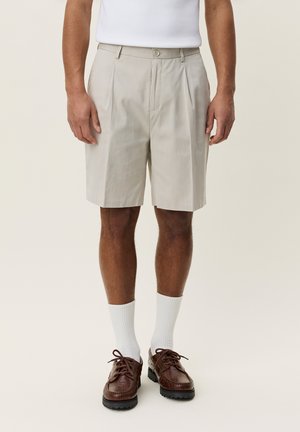 Mand iført beige plisserede shorts, hvide sokker og brune læder snørestøvler, der står mod en ensfarvet lys baggrund.