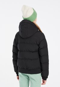 Giacca a vento nera con cappuccio, caratterizzata da un design trapuntato e polsini a coste. Indossata con pantaloni verde menta e un berretto lavorato a maglia bicolore.
