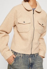 Beige fleecejakke med krave, to forlommer med klap og en frontlynlås. Struktureret materiale og cropped design, kombineret med denim.