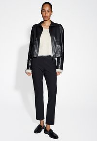 Schwarze Lederjacke in cropped Form mit Reißverschluss und zwei vorderen Taschen, kombiniert mit schwarzen, taillierten Hosen und einem weißen, flauschigen Oberteil.