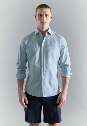 Joven con una camisa azul clara texturizada abotonada con las mangas remangadas y pantalones cortos azul marino oscuro, de pie frente a un fondo liso.