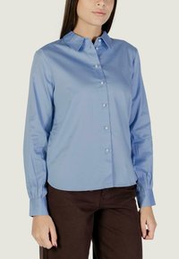 Donna che indossa una camicia azzurro chiaro a maniche lunghe con bottoni simili a perle e pantaloni marrone scuro, in piedi di fronte a uno sfondo semplice.