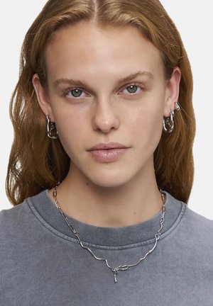Sølv kædeøreringe og et bølget sølv halskæde på en model, parret med en grå sweatshirt. Smykkerne har et moderne, abstrakt design.