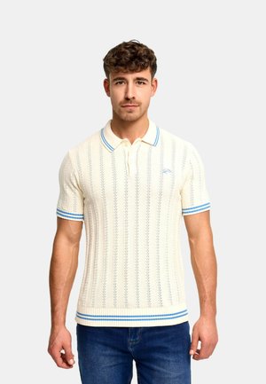 Ung mand med kort krøllet hår iført en cremefarvet strikket polo med blå striber og mørkeblå jeans, stående mod en ensartet lys baggrund.