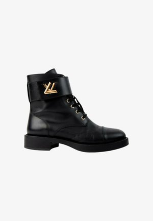 Bottine en cuir noir avec laçage devant et large sangle en haut comportant un emblème décoratif doré sur le côté extérieur.