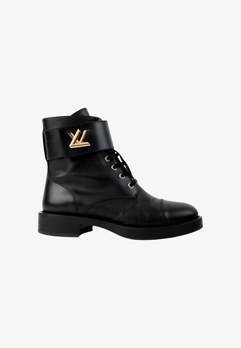 Bottine en cuir noir avec laçage devant et large sangle en haut comportant un emblème décoratif doré sur le côté extérieur.