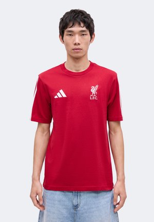 Joven con una camiseta roja del Liverpool FC Adidas y vaqueros azul claro de pie frente a un fondo liso.