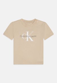 Beige puuvillainen t-paita, jossa on lyhyet hihat, resorinen pääntie ja olkapään neppari. Etupuolella on "CK"-logo ja "Calvin Klein Jeans" -teksti kontrastivärein.