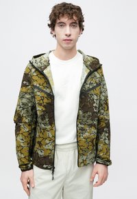 Ζακέτα παραλλαγής σε αποχρώσεις πράσινου και καφέ, με κουκούλα, κλείσιμο με φερμουάρ και ελαστικά μανίκια. Φορεμένη πάνω από λευκό φούτερ.