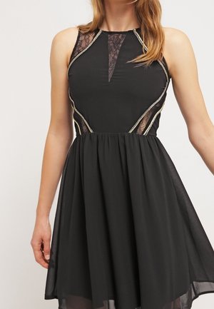Mujer con vestido negro sin mangas con paneles de encaje y ribete plateado, con cuerpo ajustado y falda fluida.