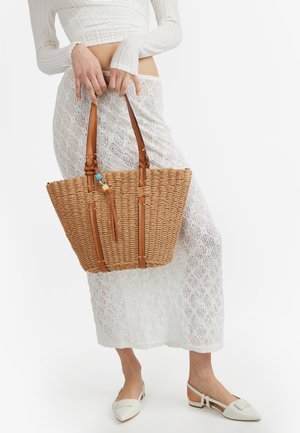 Donna con gonna e top in pizzo bianco che tiene una grande borsa intrecciata beige con manici in pelle, indossa ballerine bianche appuntite.