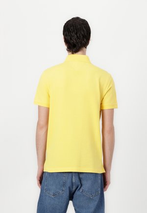 Mężczyzna w żółtej polo z krótkim rękawem i niebieskich dżinsach, stoi tyłem do widza na jednolitym białym tle.