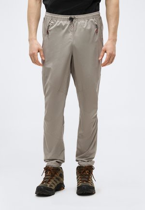 Mężczyzna w beżowych, lekkich spodniach turystycznych z elastycznym pasem i brązowych butach trekkingowych, stojący na jednolitym tle.