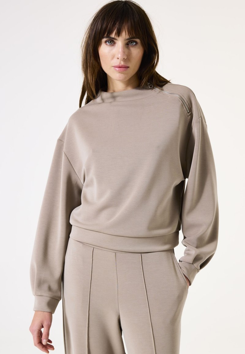 Beige Sweatshirt mit hochgeschlossenem Kragen, gerippten Bündchen und seitlichem Reißverschlusdetail. Weicher Stoff, lockere Passform, kombiniert mit passenden, taillierten Hosen.