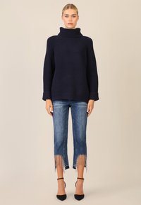 Maglione a collo alto in maglia navy con texture a costine, abbinato a jeans in denim di lavaggio medio con orli frangiati e dettagli metallici sul fondo.