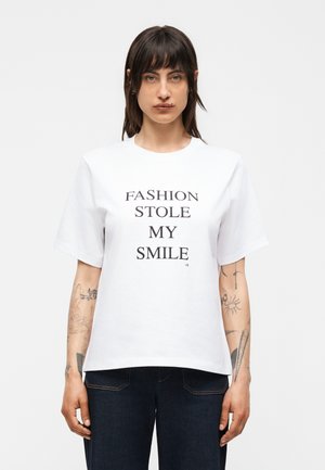 Άτομο με τατουάζ που φοράει λευκό μπλουζάκι με μαύρο κείμενο "FASHION STOLE MY SMILE" και σκούρο τζιν, κοιτάζοντας μπροστά με μονόχρωμο φόντο.