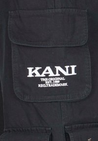 Svart stoffjakke med en klafflomme foran. "KANI"-logo i hvit tråd over lommen, sammen med teksten "THE ORIGINAL EST. 1989".
