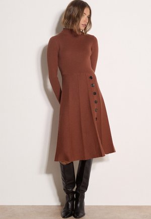 Mujer que lleva un vestido de punto de manga larga color óxido con botones laterales, combinado con botas de cuero negras hasta la rodilla, de pie frente a una pared lisa.