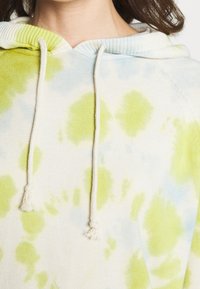 Huvtröja i tie-dye-mönster med gula och ljusblå stänk på en krämfärgad bakgrund; har dragsko med knutade ändar.