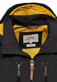 Sort jakke med en gul mesh-foring, frontlynlås med orange detaljer og et mærke, der angiver, at den er lavet af genbrugsmaterialer.