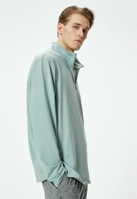 Suéter de manga larga verde claro con media cremallera y tejido suave y liso; el modelo lleva pantalones grises y está de pie de lado.