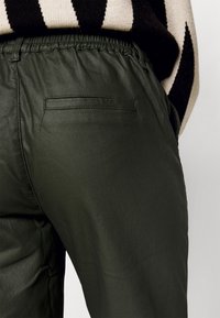 Gros plan sur un pantalon en cuir vert foncé avec poche arrière passepoilée et taille élastique, porté avec un pull rayé noir et beige.