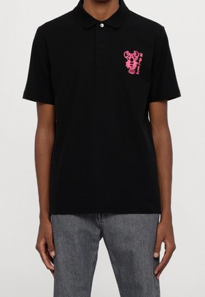 Black short-sleeve polo shirt with a pink lobster graphic and text on the left chest worn with gray jeans on a person.