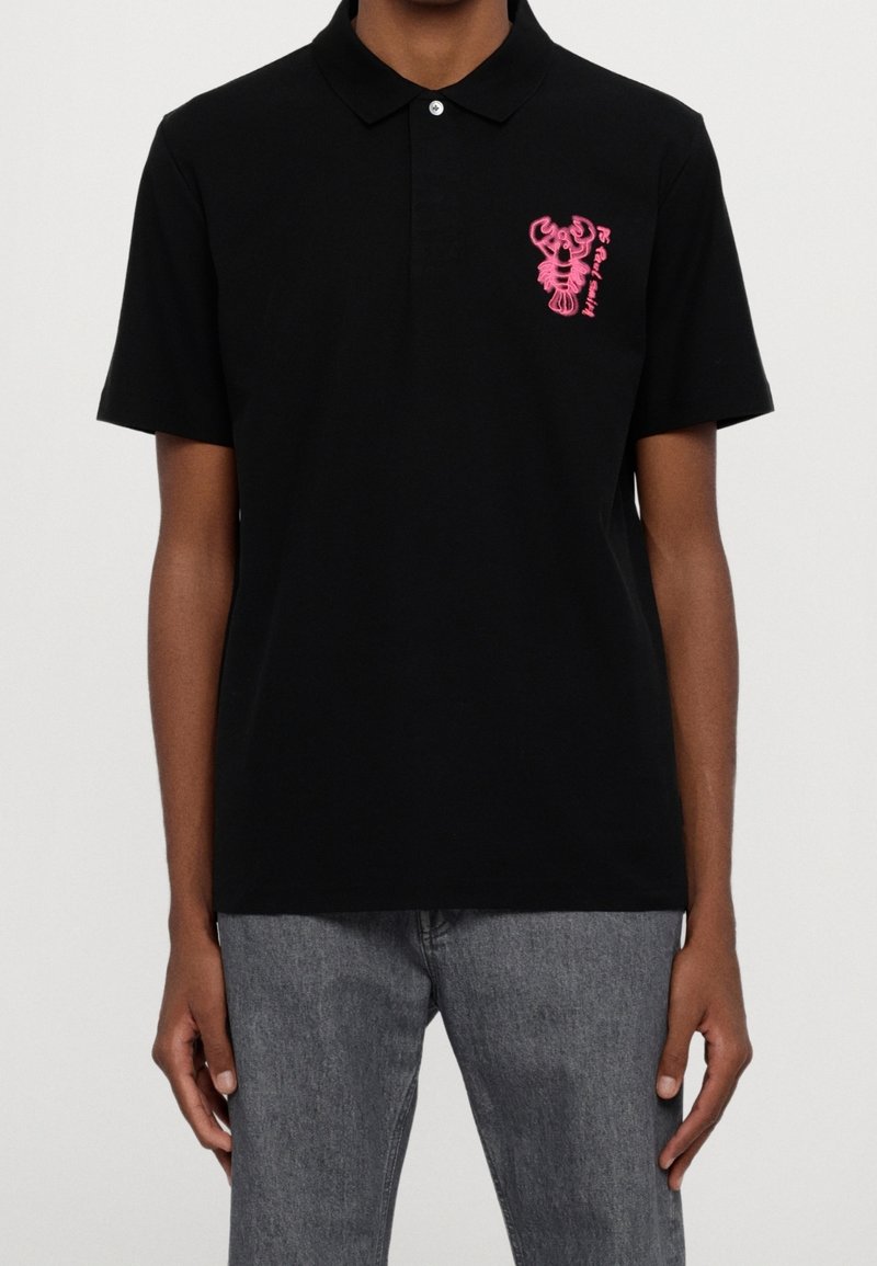 Polo noir à manches courtes avec un graphisme homard rose et du texte sur la poitrine gauche, porté avec un jean gris sur une personne.